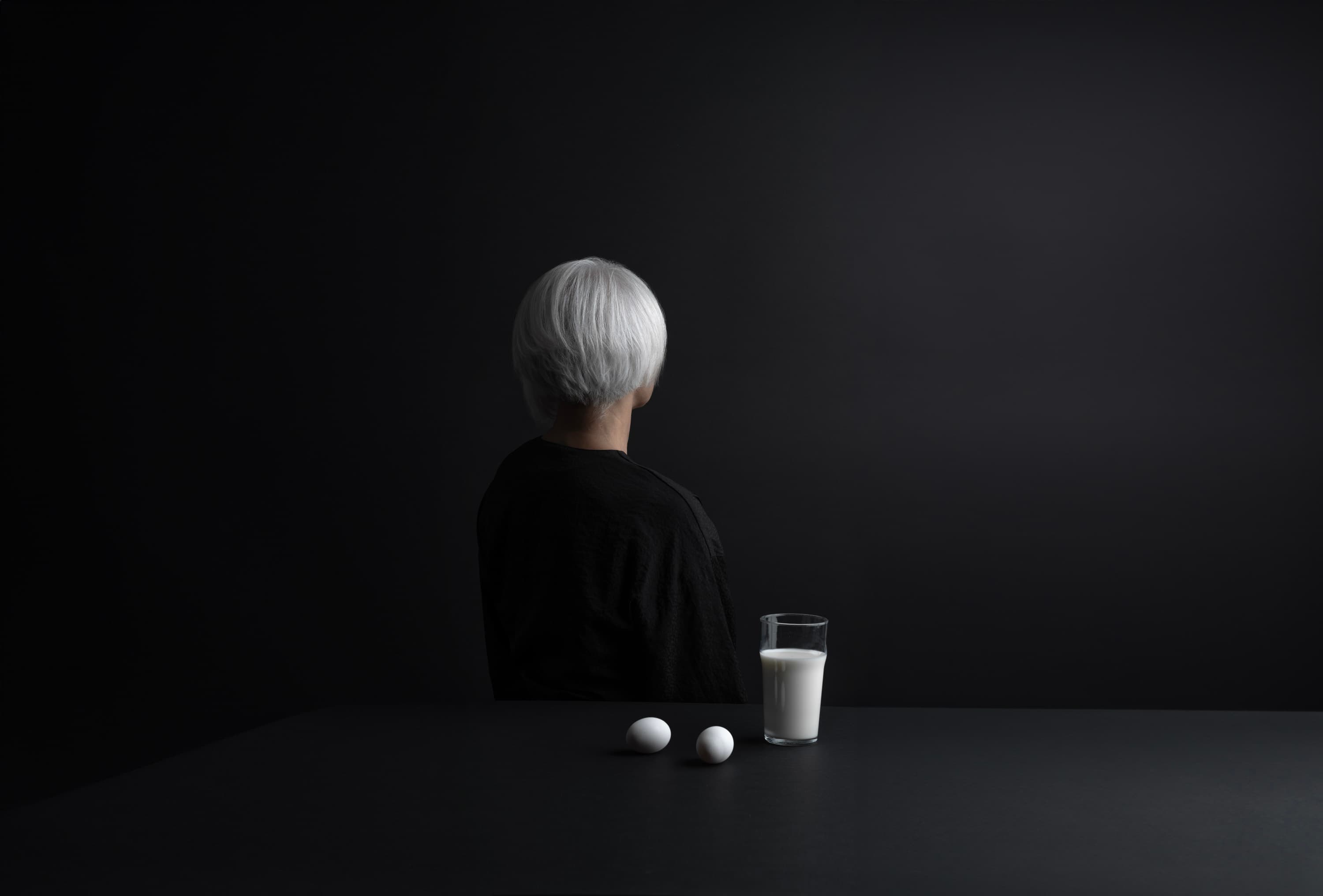 A silver-haired figure at a dark table with white objects