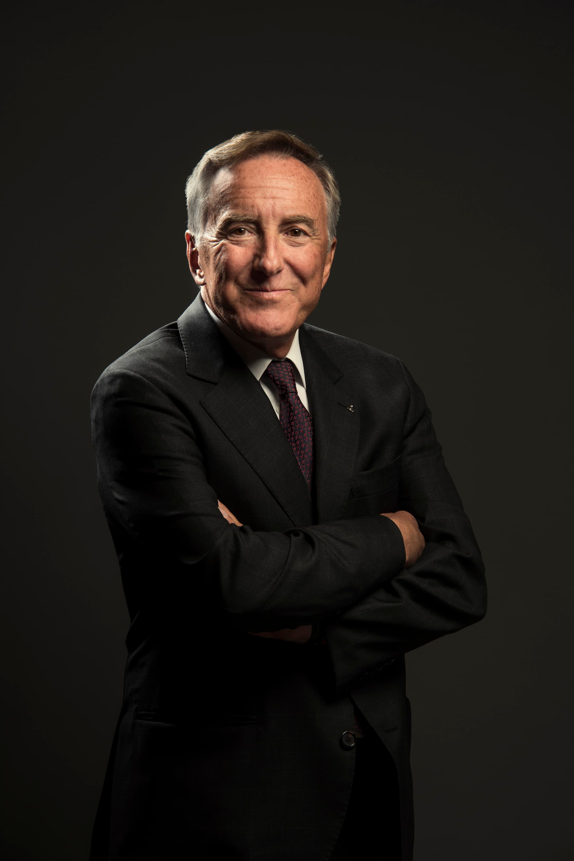 A man in a dark suit stands with his arms crossed against a dark studio backdrop