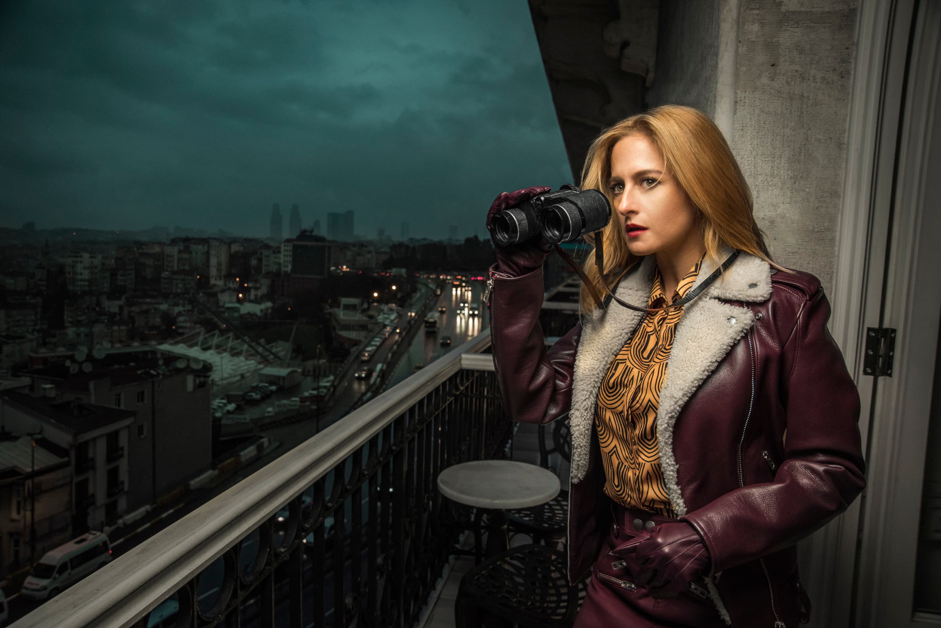 A woman in a shearling-collared leather coat looks through binoculars from a city balcony at dusk