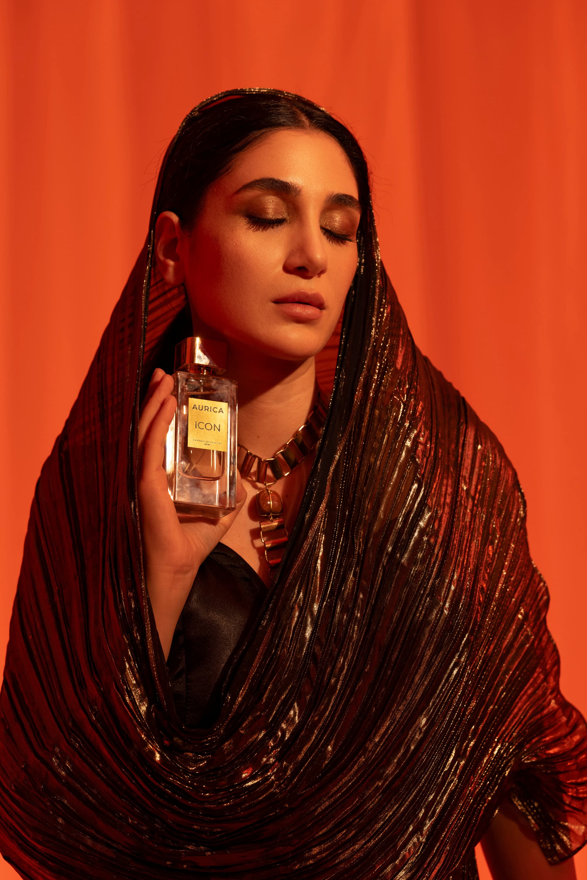 A woman in a shimmering hooded shawl holds a perfume bottle against an orange backdrop