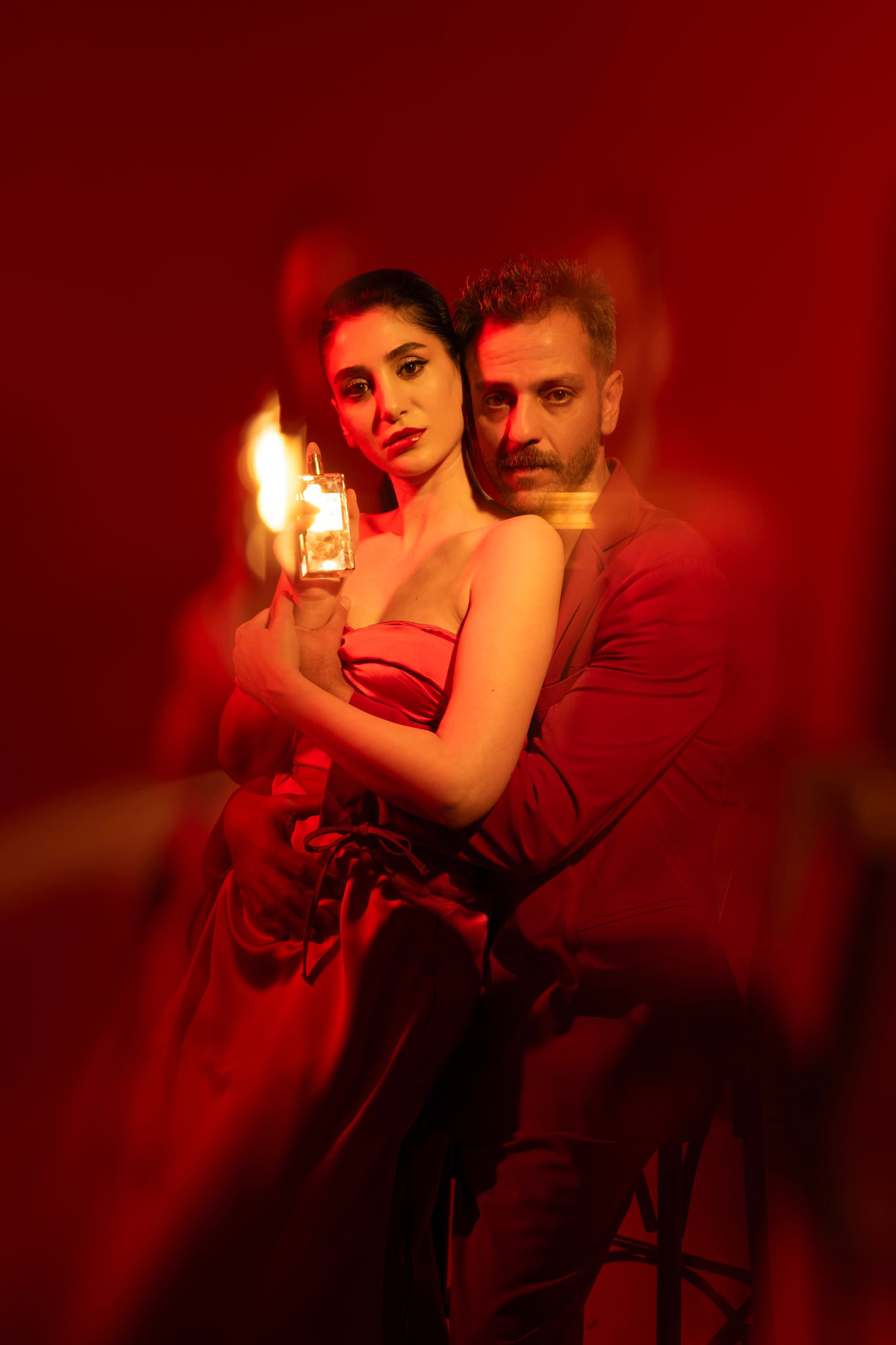 A woman holding a perfume bottle with a man embracing her from behind in red light